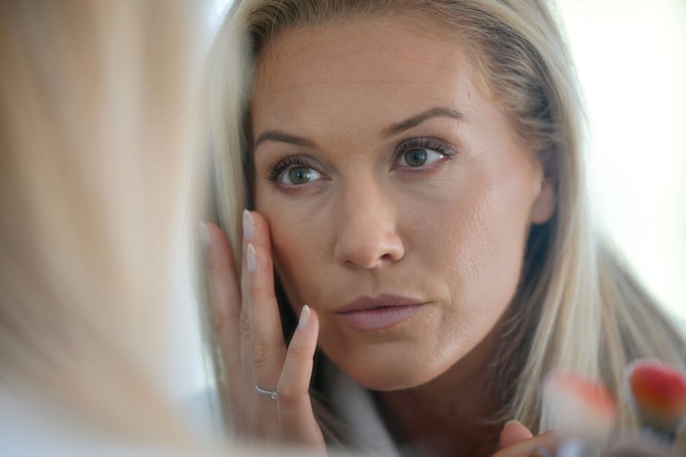 A woman looking in the mirror and considering differences between upper and lower eyelid surgery