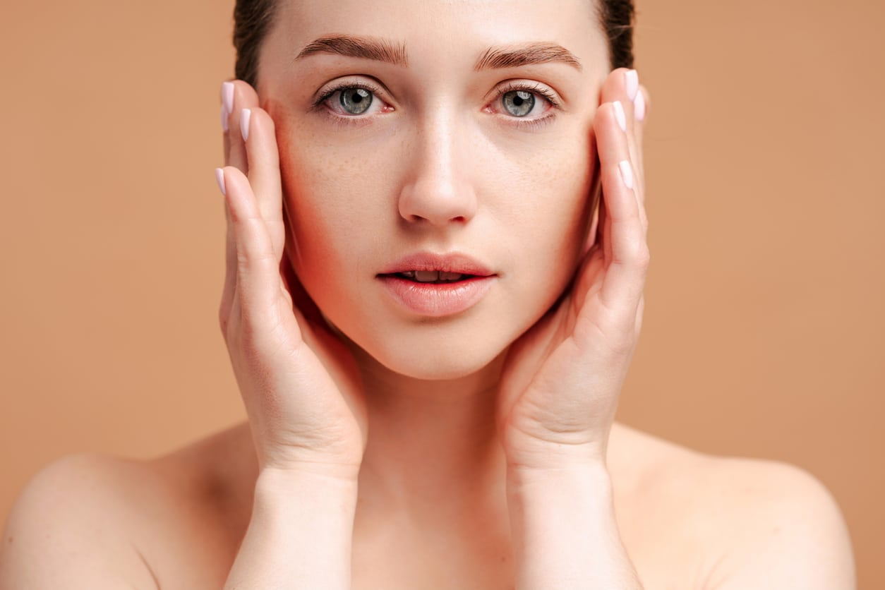 A woman looking in the mirror and considering differences between upper and lower eyelid surgery Woman touching her face with visible redness on one cheek, illustrating skin concerns treated with IPL photofacial and laser resurfacing.